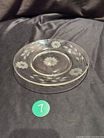 Clear glass plate with etched floral Cornflower pattern on a dark cloth background, showing full front top view.