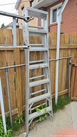 Aluminum ladder leaning against a wooden fence next to a brick wall, showing the structure and rungs of the ladder.