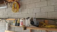 Wide shot of shelf mounted on concrete block wall with automotive fluids bottles, hardware bags, coiled yellow and red wires, and a black mechanical component.