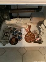 Various vintage decorative items grouped including figurines, clock parts, a tray, and other decor pieces on tiled floor under pipes.