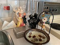 Photo showing assorted vintage decorative items including figurines of couple dancing, black torso decor piece, silver metal box, ceramic plate with floral motifs, framed clock with train motif, clear glass candlestick, and small ceramic child figurine