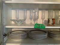 Two shelves containing assorted vintage dishes and glassware, showing stacks of ceramic plates and crystal glasses alongside decorative mugs and bowls.