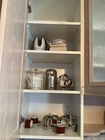 Three shelves with various vintage household dishes including cups, saucers, pitcher, canister, and decorative teapot with cups.