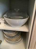 Large white bowl with glass lid on top shelf, with assorted plates stacked below on bottom shelf