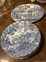 Top view of six stacked 7-inch dessert plates with blue and white pastoral patterns, and larger 10-inch plate behind them showing detailed countryside scene including people and animals.