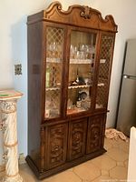 Full view of the vintage wood China cabinet showing three glass doors with gold lattice accents and three carved lower cabinet doors, with a decorative carved crest on the top.