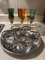 Photo showing four tall coloured glasses (green, amber, yellow) with gold rims, six clear crème brûlée dishes with silver rims arranged on a round silver tray.