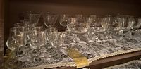 Wide view of multiple glass stemware pieces arranged on a lace doily showing various types and sizes of vintage glasses.