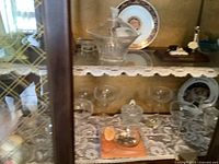 Wide shot of two shelves inside a wooden cabinet with various crystal and glass items including a decanter, glasses, covered bowls, and decorative plate