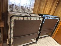Two vintage single bed cots folded and standing side by side on a tiled floor in front of a wood-paneled wall and lace curtain.