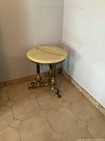 Side view of vintage Italian style marble top accent table showing round cream marble top and ornate brass base with three legs ending in curled feet.