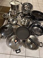 Photo showing large assortment of pots and pans of various sizes and finishes arranged on tile floor including stainless steel stock pots, frying pans, and cast iron skillet.