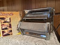 Front of metal pasta maker with hand crank beside original box