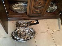 Photo showing two crystal serving platters placed on a wooden cabinet shelf, with multiple silverplated serving trays and utensils arranged on the floor beneath.