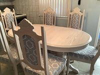 Full view of the bleached wood oval dining table with 6 matching chairs featuring carved backs and patterned silk upholstery.