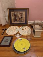 Wide view of the lot showing framed Egyptian papyrus art, floral plaques, crucifix, angel figurine, several plates and a glass lamp on a table