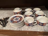 Full set of 6 espresso cups, 6 matching saucers, and 1 covered sugar bowl arranged on lace tablecloth alongside some spoons.