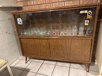 Front view of the vintage wooden storage cabinet showing glass sliding doors with glassware inside and wooden doors below, placed against a brick wall.