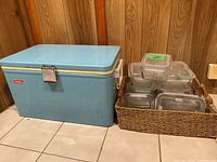 Blue Coleman cooler next to wicker basket filled with glass storage containers with lids