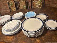 Vintage dishes arranged in groups by pattern and size on table with brick wall in background. Includes bistro set with blue rim plates, floral patterned plates, smaller saucer plates and plain white plates.