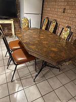 Full view of octagonal vintage kitchen table surrounded by six chairs with floral upholstered backs and brown vinyl vinyl seats, placed on tiled floor.