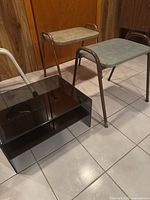Two vintage tubular metal frame stools with worn grey vinyl seats next to a black acrylic two-tier stand on a tiled floor against a wood panel wall.