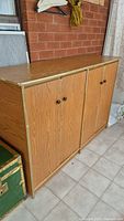 Front and side angle view of the vintage wooden storage cabinet showing three paneled doors and dark round knobs, placed against a brick wall on tiled floor.