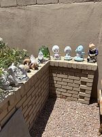 11 multi-color garden statues and figurines arranged on a brick ledge outdoors, showing various animal and gnome figures in small to medium sizes.