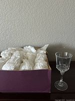 Box containing 12 crystal wine glasses individually wrapped in white tissue paper, 11 wrapped and 1 displayed outside the box showing clear, faceted design.