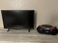 Black 24-inch Vizio TV with thin bezel and angled legs on a wood surface, next to a red and black CD player with speakers and digital display