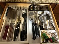 Top-down view of two white drawer organizers filled with assorted kitchen utensils including ice cream scoops, graters, potato mashers, scissors, strainers, and peelers arranged neatly in kitchen drawer under countertop.