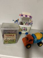 Toy ice cream cart, orange and blue cement mixer truck, and plastic bin labeled Terra with assorted dinosaur figures inside