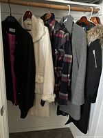 Photo showing seven coats and jackets hanging in a closet rack including black, cream with faux fur trim, plaid, gray, and black fur hooded styles.