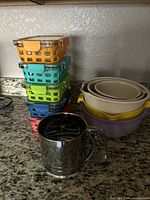 Side view of stacked silicone-covered glass containers in various colors, nested plastic bowls, and a metal sifter in front.