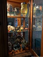 Wooden display cabinet with shelves showing multiple decorative figurines including ceramic owls, colorful glass animals, and other collectibles.