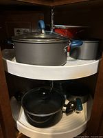 Top shelf with large and medium metal pots with blue plastic handles and glass lid, black metal pot with glass lid and black handles below