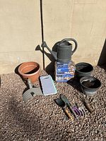 Photo showing assembly of gardening tools including watering can, shovels, hand rake, pruners, knee pad, and plastic pots situated on gravel against a wall.