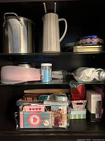 Metal pot, ceramic pitcher and cookie tins on upper shelf. Various baking supplies and cotton candy maker on lower shelves.