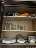Kitchen cabinet with three shelves showing stacked ceramic plates, bowls, and glass cups on two shelves.