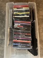 Top-down view of a plastic bin filled with various music CDs including Eminem, Savage Garden, Enrique Iglesias, and others showing titles and cases.