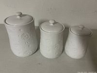 Front view of three white ceramic canisters of varying sizes, all with embossed floral designs and fitted lids with knobs.