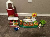 Full set arranged on the floor showing the red barn, farm stand with roof, green tractor, base platform with fence and flower, dog and cow Little People figures, blue cart, and farmer figure.