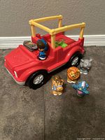 Photo showing the red and yellow Safari Jeep toy with the girl figure sitting in the driver's seat and four animal figures (elephant, lion, bird, and a smaller animal figure) arranged in front of the Jeep on the floor.