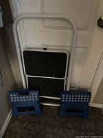 Photo showing white folding step ladder closed with two blue plastic step stools placed in front, near a white door.
