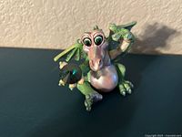 Front view of the figurine showing the dragon sitting, green color with pink belly and large crystal eyes holding a crystal orb.