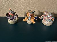 Set of three Franklin Mint Mood Dragons figurines displayed on a dark surface against wall, viewed from front. Dragons are sitting on cracked cream eggs.