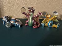Three Franklin Mint Mood Dragons figurines displayed side by side on a flat surface against a wall. Each dragon is a different color with detailed features and crystal accents, holding a clear glass orb.