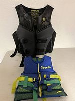 Front view of black adult large Hyperlite life jacket and blue/green child Speedo life jacket on table