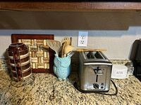 Photo showing stainless steel two-slice toaster, wooden utensils in light blue owl mug, and ceramic utensil holder with tile pattern placed on kitchen counter.