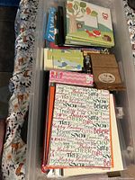 Clear plastic container filled with scrapbooking supplies including albums with holiday and animal themes, embellishments, and decorative papers.